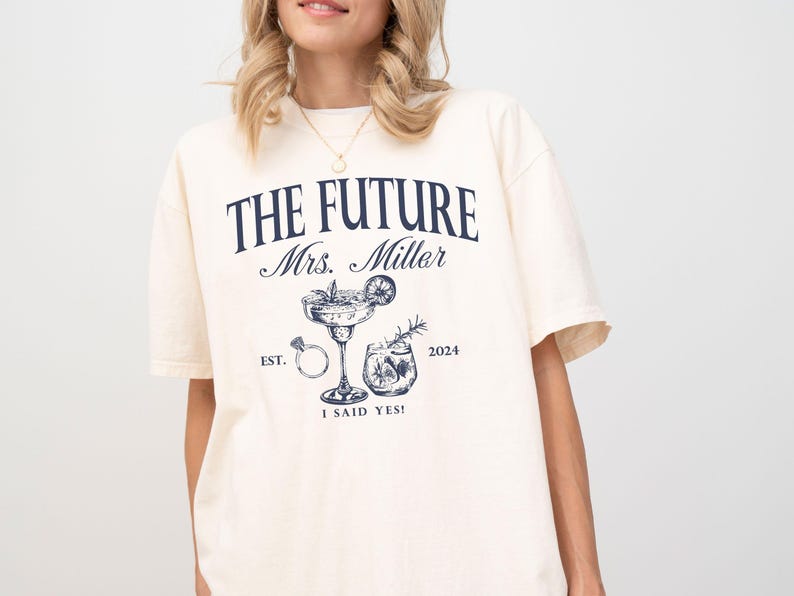 May include: A white t-shirt with the text "The Future Mrs. Miller Est. 2024 I Said Yes!" printed on it. The design also includes a cocktail glass with a ring and a small glass of liquid.