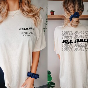 K&ouml;nnte beinhalten: Ein wei&szlig;es T-Shirt mit dem Text "MRS. JAMES" in Schwarz auf der Vorderseite und dem gleichen Text f&uuml;nfmal in einem gestapelten Muster auf der R&uuml;ckseite. Der Text ist in einem Retro-Schriftart-Stil.