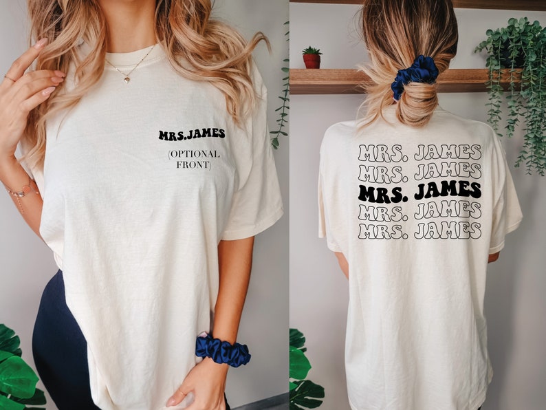 K&ouml;nnte beinhalten: Ein wei&szlig;es T-Shirt mit dem Text "MRS. JAMES" in Schwarz auf der Vorderseite und dem gleichen Text f&uuml;nfmal in einem gestapelten Muster auf der R&uuml;ckseite. Der Text ist in einem Retro-Schriftart-Stil.