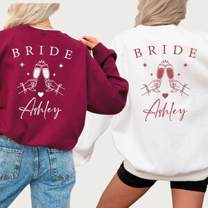 May include: Two sweatshirts, one burgundy and one white, feature the word "BRIDE" above a graphic of two champagne flutes clinking. Below the graphic is the name "Ashley" in script. The burgundy sweatshirt is paired with blue jeans.