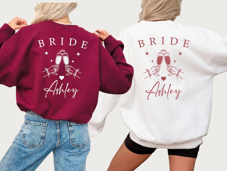 May include: Two sweatshirts, one burgundy and one white, feature the word "BRIDE" above a graphic of two champagne flutes clinking. Below the graphic is the name "Ashley" in script. The burgundy sweatshirt is paired with blue jeans.
