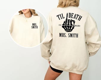 May include: A beige sweatshirt with the text "TIL DEATH EST. 01.04.2024 MRS. SMITH" and a black skeleton hand giving the middle finger on the back. The front of the sweatshirt has the text "MRS. SMITH" in black.