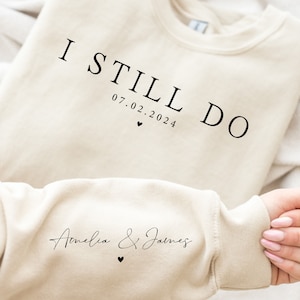 May include: A beige sweatshirt with the text "I Still Do 07.02.2024" on the front and "Amelia & James" on the sleeve.