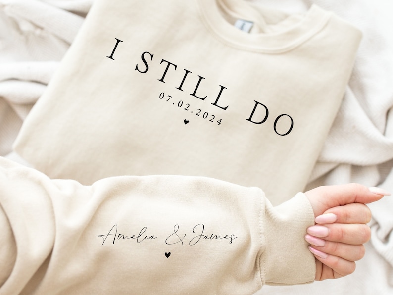 May include: A beige sweatshirt with the text "I Still Do 07.02.2024" on the front and "Amelia & James" on the sleeve.