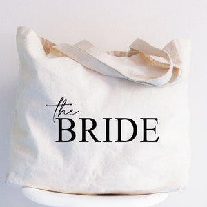 May include: An off-white canvas tote bag featuring two fabric handles and the black printed text "the BRIDE" on the front. The word "the" is written in a cursive script font, while the word "BRIDE" is in a bold, uppercase serif font.