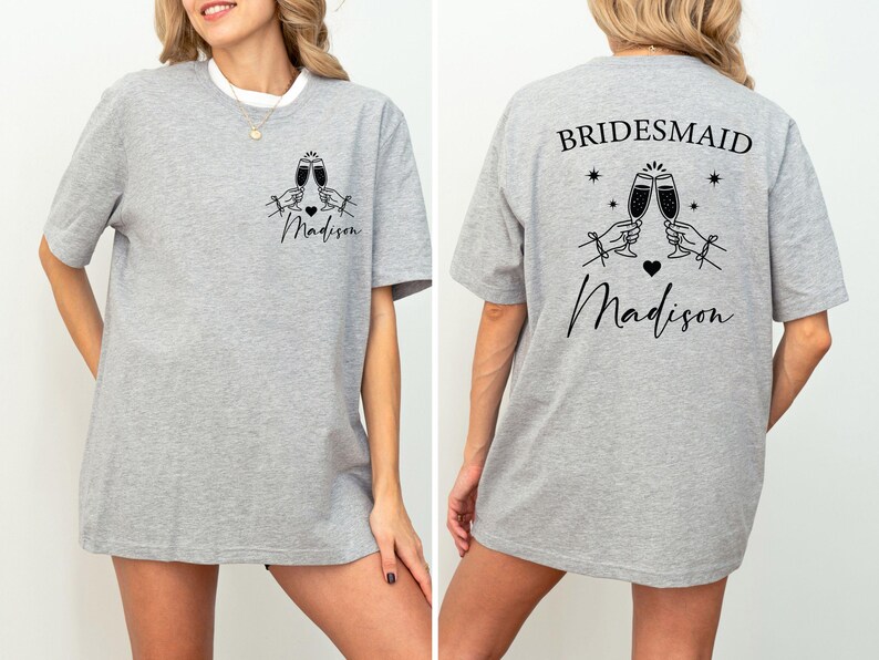 May include: A light gray t-shirt with a white collar. The front features a graphic of two champagne glasses clinking, a heart, and the name "Madison". The back says "Bridesmaid" above the same graphic and name.