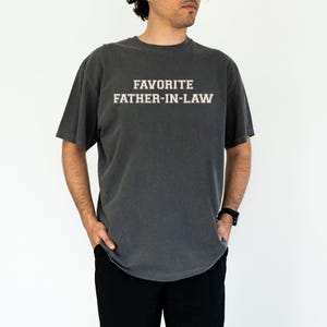 May include: Dark gray t-shirt with the text "FAVORITE FATHER-IN-LAW" in white block letters. The shirt has short sleeves and a crew neck. The model is wearing black pants and a black watch.