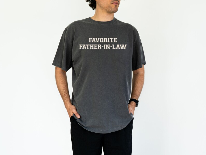 May include: Dark gray t-shirt with the text "FAVORITE FATHER-IN-LAW" in white block letters. The shirt has short sleeves and a crew neck. The model is wearing black pants and a black watch.
