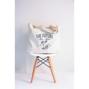 K&ouml;nnte beinhalten: Eine cremefarbene Tragetasche mit dem Text "THE FUTURE Mrs. Miller" in Marineblau und einer Cocktail-Illustration. Die Tasche steht auf einem wei&szlig;en Stuhl mit Holzbeinen. Die Tasche hat einen Griff.
