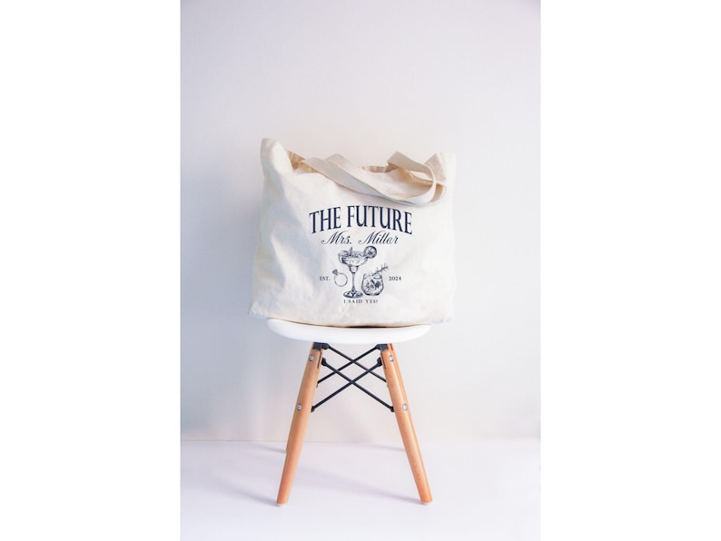 K&ouml;nnte beinhalten: Eine cremefarbene Tragetasche mit dem Text "THE FUTURE Mrs. Miller" in Marineblau und einer Cocktail-Illustration. Die Tasche steht auf einem wei&szlig;en Stuhl mit Holzbeinen. Die Tasche hat einen Griff.
