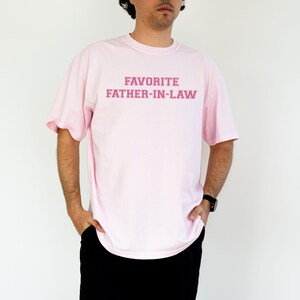 May include: A light pink t-shirt with the text "FAVORITE FATHER-IN-LAW" in a darker pink font. The shirt is a crew neck style and appears to be made of a soft material. The person is wearing black pants and a black watch.
