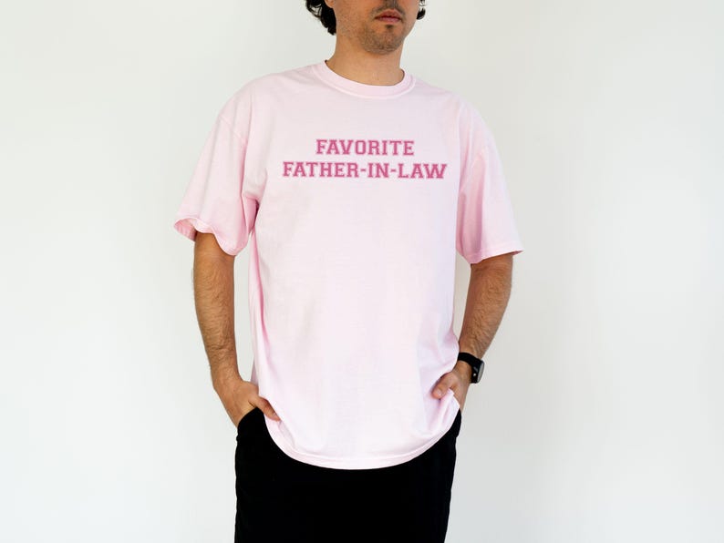 May include: A light pink t-shirt with the text "FAVORITE FATHER-IN-LAW" in a darker pink font. The shirt is a crew neck style and appears to be made of a soft material. The person is wearing black pants and a black watch.