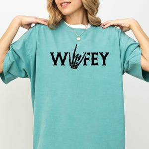 May include: A turquoise t-shirt with the word "Wifey" in black gothic lettering, featuring a skeleton hand making a gesture. The shirt has short sleeves and a relaxed fit.