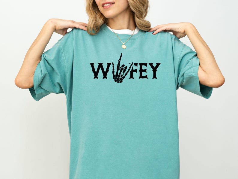 May include: A turquoise t-shirt with the word "Wifey" in black gothic lettering, featuring a skeleton hand making a gesture. The shirt has short sleeves and a relaxed fit.