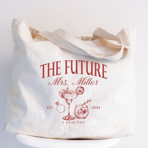 K&ouml;nnte beinhalten: Beige Canvas-Tragetasche mit rotem Text und Illustrationen. Der Text lautet "THE FUTURE Mrs. Miller" &uuml;ber einer Zeichnung eines Cocktails, eines Rings und anderer Getr&auml;nke. Der Text "EST. 2024" und "I SAID YES!" sind ebenfalls enthalten.