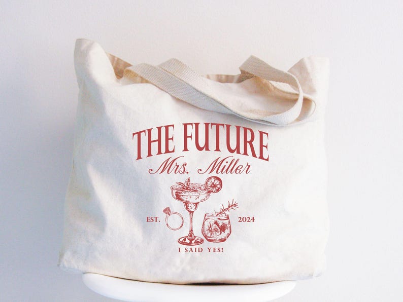 K&ouml;nnte beinhalten: Beige Canvas-Tragetasche mit rotem Text und Illustrationen. Der Text lautet "THE FUTURE Mrs. Miller" &uuml;ber einer Zeichnung eines Cocktails, eines Rings und anderer Getr&auml;nke. Der Text "EST. 2024" und "I SAID YES!" sind ebenfalls enthalten.