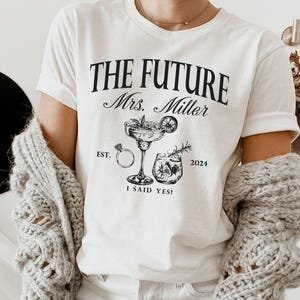 May include: White t-shirt with black text that reads "The Future Mrs. Miller Est. 2024 I Said Yes!" and a graphic of a cocktail glass and a ring.