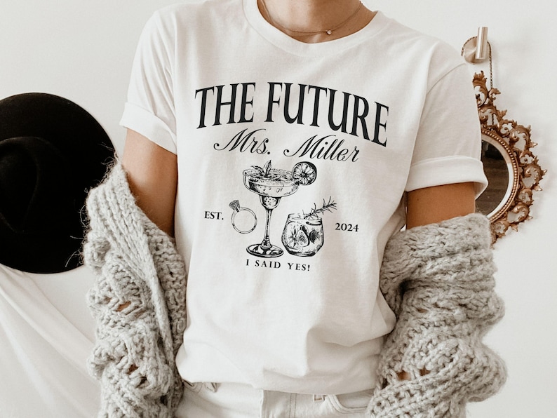 May include: White t-shirt with black text that reads "The Future Mrs. Miller Est. 2024 I Said Yes!" and a graphic of a cocktail glass and a ring.