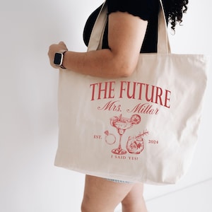 K&ouml;nnte beinhalten: Cremefarbene Tragetasche mit rotem Text und Illustrationen. Der Text lautet "THE FUTURE Mrs. Miller" &uuml;ber einem Cocktailglas, einem Ring und anderen Illustrationen. Der untere Text lautet "I SAID YES!" und "2024".