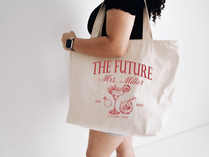K&ouml;nnte beinhalten: Cremefarbene Tragetasche mit rotem Text und Illustrationen. Der Text lautet "THE FUTURE Mrs. Miller" &uuml;ber einem Cocktailglas, einem Ring und anderen Illustrationen. Der untere Text lautet "I SAID YES!" und "2024".