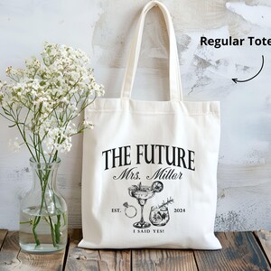 K&ouml;nnte beinhalten: Cremefarbene Tragetasche mit dem Text "THE FUTURE Mrs. Miller" und dem Jahr 2024. Die Tasche zeigt Illustrationen eines Cocktails, eines Rings und eines Getr&auml;nks. Die Worte "I SAID YES!" sind ebenfalls auf der Tasche aufgedruckt.