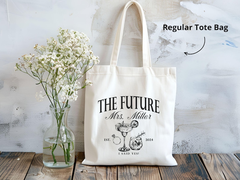 K&ouml;nnte beinhalten: Cremefarbene Tragetasche mit dem Text "THE FUTURE Mrs. Miller" und dem Jahr 2024. Die Tasche zeigt Illustrationen eines Cocktails, eines Rings und eines Getr&auml;nks. Die Worte "I SAID YES!" sind ebenfalls auf der Tasche aufgedruckt.