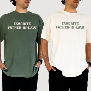 May include: Two short-sleeved t-shirts, one olive green and one cream, each with the text "FAVORITE FATHER-IN-LAW" in a block font. The shirts are paired with black pants. The shirts appear to be made of a soft, comfortable material.