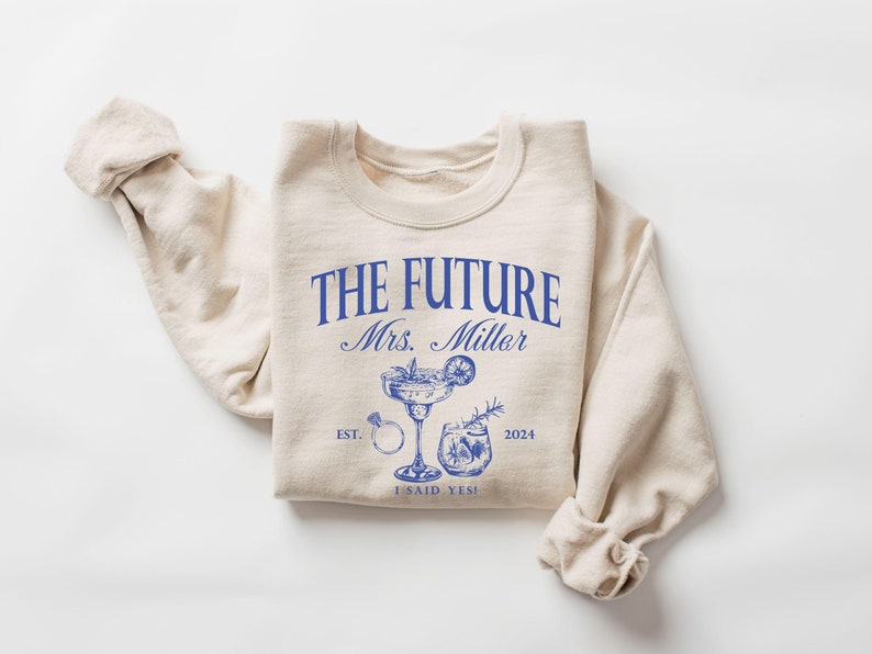May include: A beige sweatshirt with the text "THE FUTURE Mrs. Miller EST. 2024 I SAID YES!" in blue lettering. The design also includes a cocktail glass with a lime wedge and a ring.