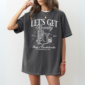May include: A dark gray t-shirt with white text that reads "Let's Get Rowdy" with a boot and a cocktail glass graphic. The text also includes "Est. 2024" and "Amy's Bachelorette Nashville, TN".