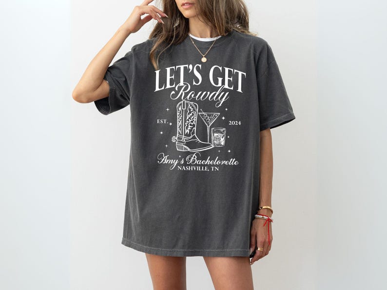 May include: A dark gray t-shirt with white text that reads "Let's Get Rowdy" with a boot and a cocktail glass graphic. The text also includes "Est. 2024" and "Amy's Bachelorette Nashville, TN".