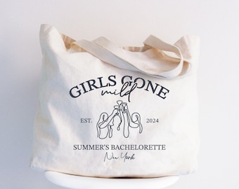 Puede incluir: Bolso tote de color crema con el texto "GIRLS GONE mild" en negro, en forma de arco. Debajo, un dibujo lineal de tres figuras brindando con copas. El texto "SUMMER'S BACHELORETTE New York" está debajo de la ilustración.