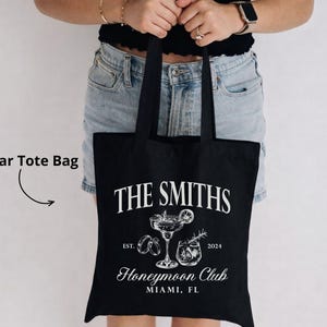 May include: Black tote bag with white text and illustrations. The text reads "THE SMITHS" above a cocktail glass and the words "Honeymoon Club Miami, FL". The bag also has the text "EST. 2024".