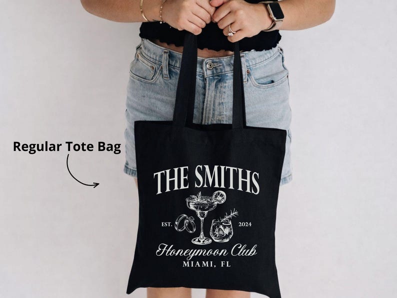 May include: Black tote bag with white text and illustrations. The text reads "THE SMITHS" above a cocktail glass and the words "Honeymoon Club Miami, FL". The bag also has the text "EST. 2024".