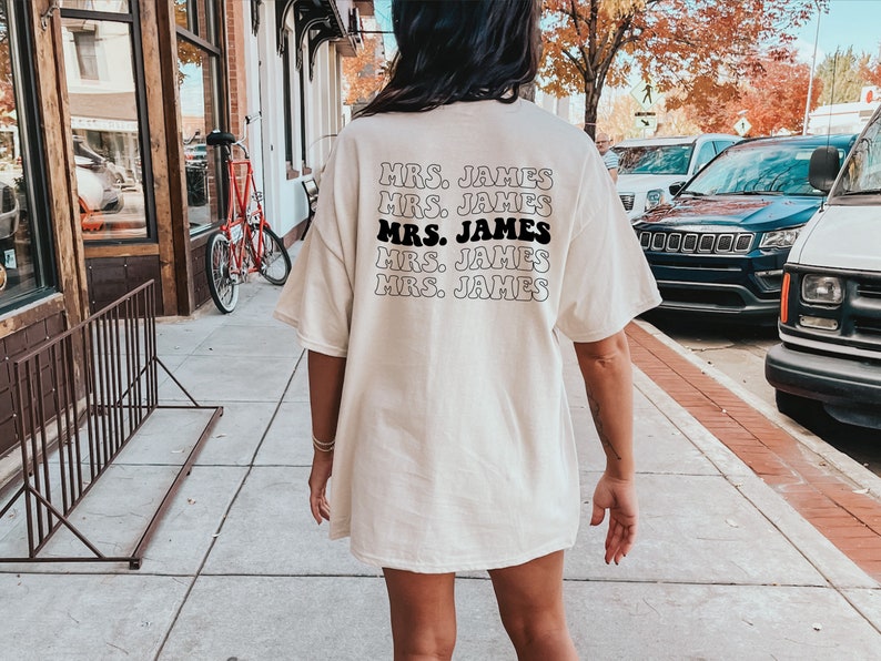 K&ouml;nnte beinhalten: Ein wei&szlig;es Oversize-T-Shirt mit dem Text "MRS. JAMES", der f&uuml;nfmal in schwarzer, Retro-Schrift wiederholt wird.