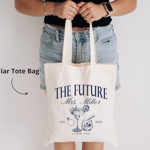 K&ouml;nnte beinhalten: Cremefarbene Tragetasche mit marineblauem Text und Illustrationen. Der Text lautet "THE FUTURE Mrs. Millar" mit "EST. 2024 I SAID YES!" darunter. Enth&auml;lt einen Cocktail, einen Ring und andere Grafiken. Die Tasche hat zwei Griffe.