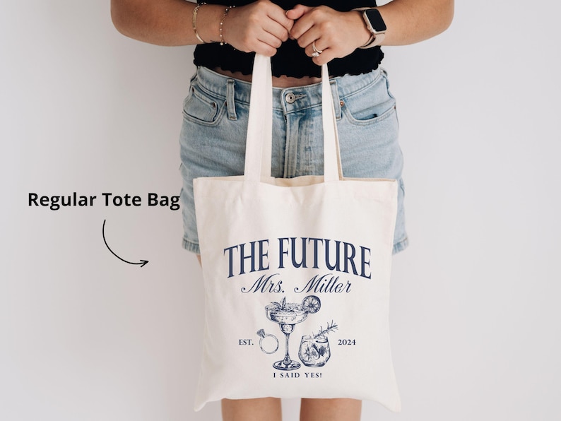 K&ouml;nnte beinhalten: Cremefarbene Tragetasche mit marineblauem Text und Illustrationen. Der Text lautet "THE FUTURE Mrs. Millar" mit "EST. 2024 I SAID YES!" darunter. Enth&auml;lt einen Cocktail, einen Ring und andere Grafiken. Die Tasche hat zwei Griffe.