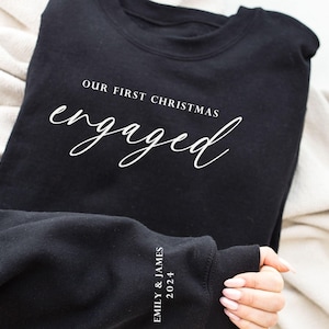 May include: Black sweatshirt with the text "OUR FIRST CHRISTMAS engaged" in white script. The sleeve displays "EMILY & JAMES 2024". A cozy, casual garment, perfect for celebrating an engagement during the holiday season.