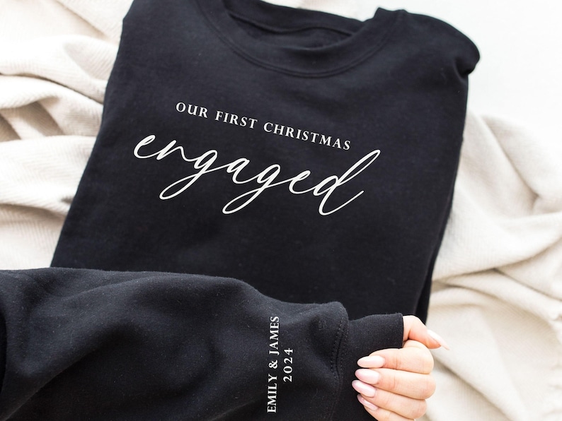 May include: Black sweatshirt with the text "OUR FIRST CHRISTMAS engaged" in white script. The sleeve displays "EMILY & JAMES 2024". A cozy, casual garment, perfect for celebrating an engagement during the holiday season.