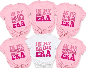 Op de afbeelding: Zes roze en witte T-shirts met de tekst "In My Bride Era" in een retro-lettertype. De shirts hebben verschillende variaties van de tekst, waaronder "Bride Squad", "Bridesmaid", "Maid of Honor", "Mother of the Bride" en "Sister of the Bride".