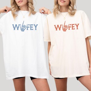 May include: Two oversized t-shirts, one white and one cream, each with the word "Wifey" printed in a stylized font. The white shirt has blue text and a skeleton hand graphic, while the cream shirt has red text and a matching graphic.
