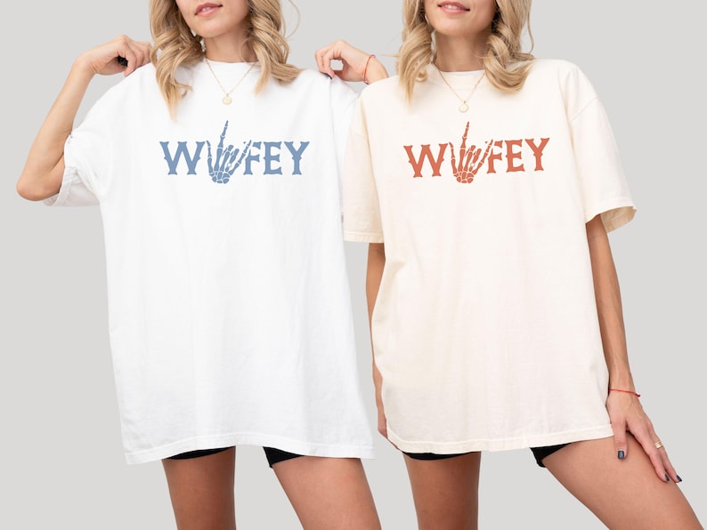 May include: Two oversized t-shirts, one white and one cream, each with the word "Wifey" printed in a stylized font. The white shirt has blue text and a skeleton hand graphic, while the cream shirt has red text and a matching graphic.