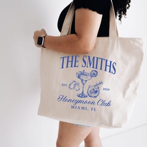 May include: Cream-colored tote bag with blue text and illustrations. The text reads "THE SMITHS" above a cocktail illustration, "Honeymoon Club" and "MIAMI, FL". The bag is held by a person.