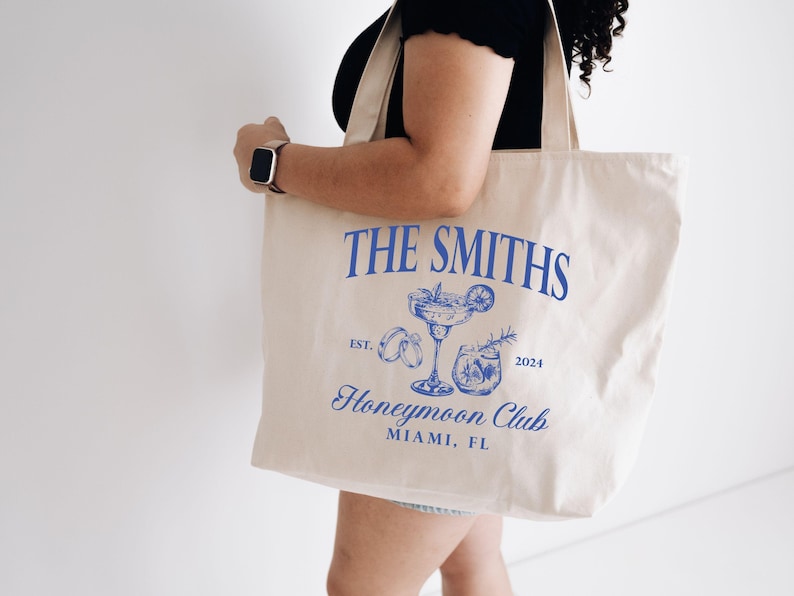May include: Cream-colored tote bag with blue text and illustrations. The text reads "THE SMITHS" above a cocktail illustration, "Honeymoon Club" and "MIAMI, FL". The bag is held by a person.