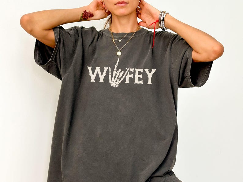 May include: Dark gray oversized t-shirt with the word "Wifey" in white, featuring a skeleton hand giving the middle finger. The shirt has a vintage, worn look.