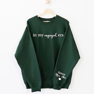 Puede incluir: Una sudadera verde oscuro con la frase "in my engaged era" impresa en blanco en el pecho. La manga presenta el texto "future Mrs. Alex" con un s&iacute;mbolo de coraz&oacute;n. La sudadera est&aacute; colgada en una percha de madera.