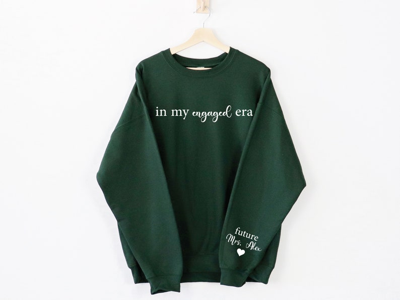 Puede incluir: Una sudadera verde oscuro con la frase "in my engaged era" impresa en blanco en el pecho. La manga presenta el texto "future Mrs. Alex" con un s&iacute;mbolo de coraz&oacute;n. La sudadera est&aacute; colgada en una percha de madera.