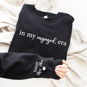 Puede incluir: Sudadera negra con la frase "in my engaged era" en escritura blanca. La manga muestra el texto "future Mrs. Alex" con un peque&ntilde;o coraz&oacute;n blanco. La sudadera est&aacute; doblada sobre una tela color crema.