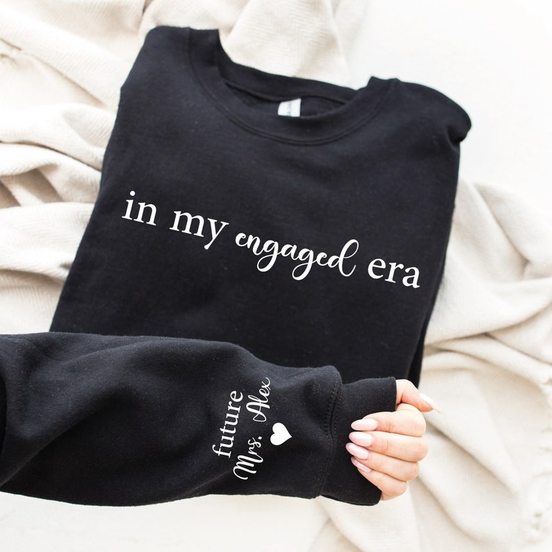 Puede incluir: Sudadera negra con la frase "in my engaged era" en escritura blanca. La manga muestra el texto "future Mrs. Alex" con un peque&ntilde;o coraz&oacute;n blanco. La sudadera est&aacute; doblada sobre una tela color crema.