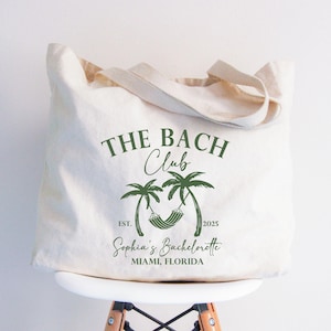 May include: Cream-colored tote bag with green text and graphics. The text reads "THE BACH Club" above a palm tree graphic with a hammock. Below the graphic is "EST. 2025" and "Sophia's Bachelorette Miami, Florida".