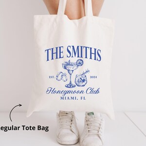 May include: Cream-colored tote bag with blue text and illustrations. The text reads "THE SMITHS Honeymoon Club Miami, FL" with the year 2024. Illustrations include a cocktail, rings, and a drink. The bag has two handles and is held up.